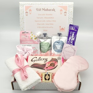 May include: A pink and white gift box filled with various items, including a white towel with a pink ribbon, a chocolate bar, a face mask, a sleep mask, and a small gold decorative item. The box has a pink and white patterned design with gold stars. The box also has a card with the text "Eid Mubarak" and a message about starting and ending with Bismillah and Alhamdulillah.