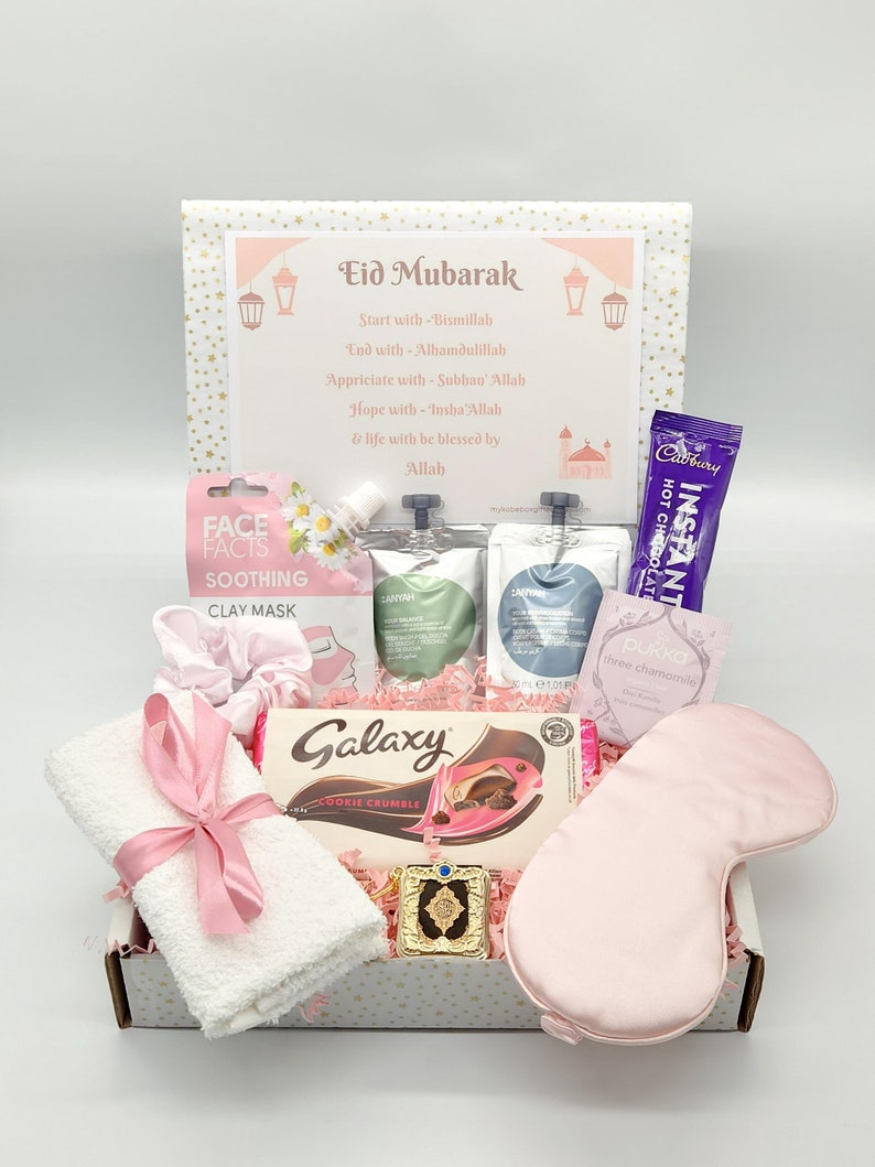 May include: A pink and white gift box filled with various items, including a white towel with a pink ribbon, a chocolate bar, a face mask, a sleep mask, and a small gold decorative item. The box has a pink and white patterned design with gold stars. The box also has a card with the text "Eid Mubarak" and a message about starting and ending with Bismillah and Alhamdulillah.