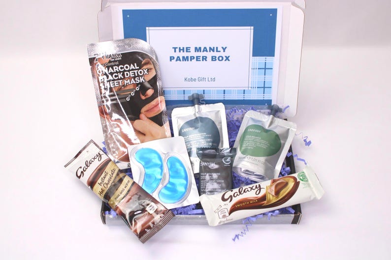 May include: A gift box labeled "THE MANLY PAMPER BOX" filled with skincare products, eye masks, and Galaxy chocolate bars. A charcoal black detox sheet mask is visible, along with other self-care items. The box is lined with purple shredded paper.