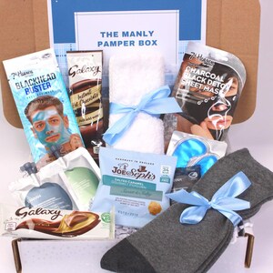 May include: A gift box labeled "THE MANLY PAMPER BOX" filled with self-care items. Includes a face mask, instant hot chocolate, a towel tied with a blue ribbon, eye masks, socks, and a chocolate bar.