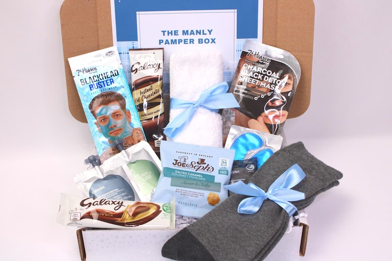 May include: A gift box labeled "THE MANLY PAMPER BOX" filled with self-care items. Includes a face mask, instant hot chocolate, a towel tied with a blue ribbon, eye masks, socks, and a chocolate bar.