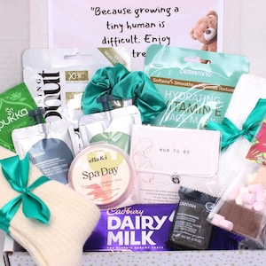 May include: A gift box filled with self-care items. Includes a green scrunchie, face mask, tea, spa day cream, socks, chocolate, and a bracelet. The box has a sign that reads "Because growing a tiny human is difficult. Enjoy tre".