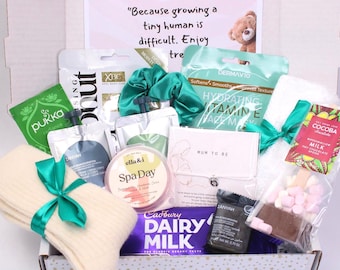 May include: A gift box filled with self-care items. Includes a green scrunchie, face mask, tea, spa day cream, socks, chocolate, and a bracelet. The box has a sign that reads "Because growing a tiny human is difficult. Enjoy tre".