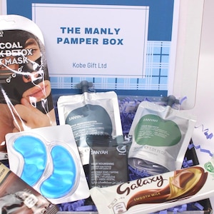 May include: A gift box labeled "THE MANLY PAMPER BOX" contains skincare products, including a charcoal sheet mask, eye patches, and various lotions. A Galaxy chocolate bar and a tube of instant hot chocolate are also included. The box is decorated with blue and white accents.