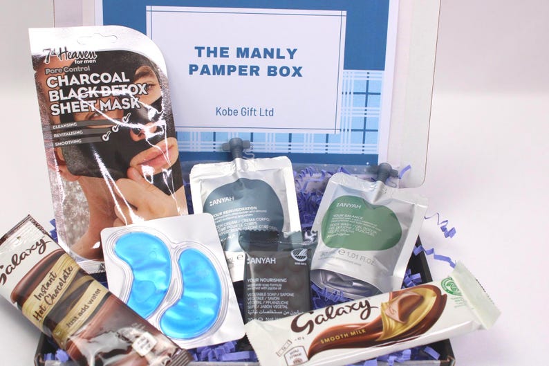 May include: A gift box labeled "THE MANLY PAMPER BOX" contains skincare products, including a charcoal sheet mask, eye patches, and various lotions. A Galaxy chocolate bar and a tube of instant hot chocolate are also included. The box is decorated with blue and white accents.