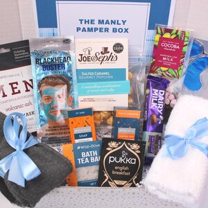 May include: A gift box labeled "THE MANLY PAMPER BOX" filled with self-care items. Includes a face mask, popcorn, chocolate, tea, a washcloth with a blue ribbon, and other grooming products. The box is white with blue accents.