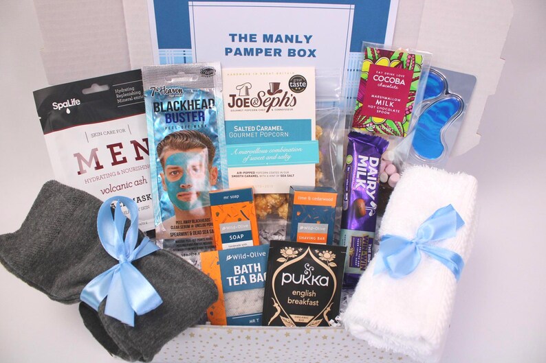 May include: A gift box labeled "THE MANLY PAMPER BOX" filled with self-care items. Includes a face mask, popcorn, chocolate, tea, a washcloth with a blue ribbon, and other grooming products. The box is white with blue accents.