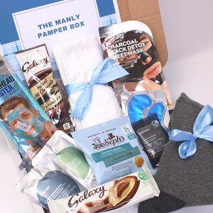 May include: A gift box labeled "The Manly Pamper Box" filled with self-care items. Includes a face mask, hot chocolate, a towel tied with a blue ribbon, eye masks, socks, and snacks. The box is brown with a blue and white label.