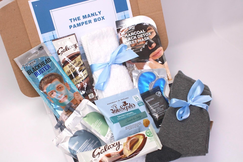 May include: A gift box labeled "The Manly Pamper Box" filled with self-care items. Includes a face mask, hot chocolate, a towel tied with a blue ribbon, eye masks, socks, and snacks. The box is brown with a blue and white label.