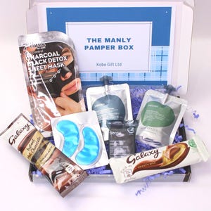May include: A gift box labeled "THE MANLY PAMPER BOX" contains skincare products, eye masks, and chocolate bars. The box includes a charcoal face mask, eye gel pads, and Galaxy chocolate bars. The box is filled with purple paper.