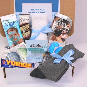 May include: A gift box labeled "THE MANLY PAMPER BOX" filled with self-care items. Includes a face mask, instant hot chocolate, a towel, eye masks, socks, and a chocolate bar. The box is tied with blue ribbons.