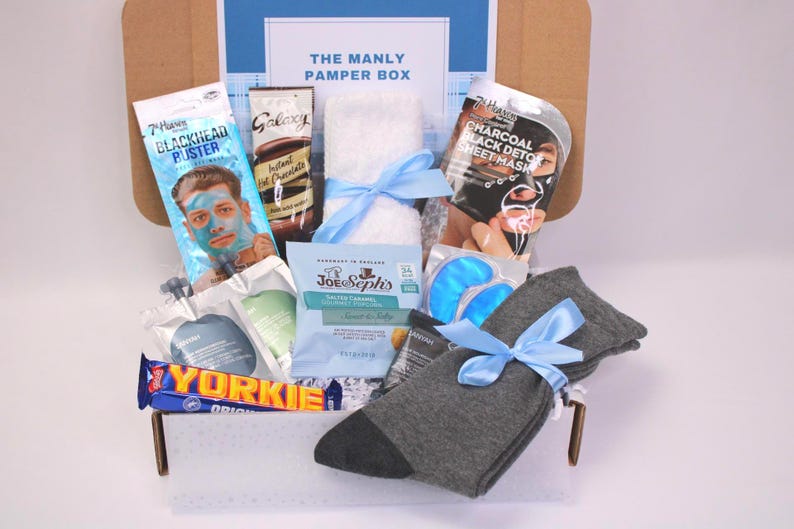 May include: A gift box labeled "THE MANLY PAMPER BOX" filled with self-care items. Includes a face mask, instant hot chocolate, a towel, eye masks, socks, and a chocolate bar. The box is tied with blue ribbons.