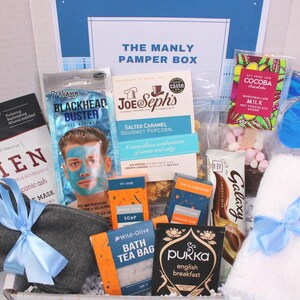 May include: A gift box labeled "THE MANLY PAMPER BOX" filled with self-care items. Includes a face mask, popcorn, chocolate, bath tea bags, a towel, and eye mask. The box is tied with a blue ribbon.
