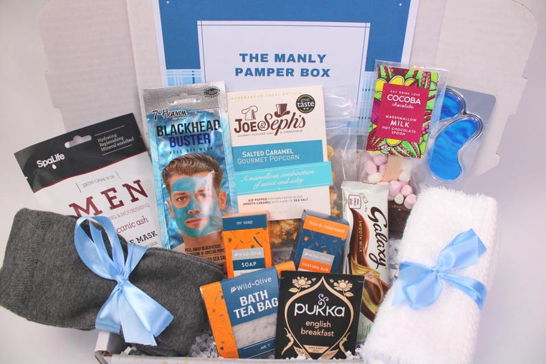 May include: A gift box labeled "THE MANLY PAMPER BOX" filled with self-care items. Includes a face mask, popcorn, chocolate, bath tea bags, a towel, and eye mask. The box is tied with a blue ribbon.