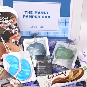 May include: A gift box labeled "THE MANLY PAMPER BOX" contains skincare products, including a charcoal sheet mask, eye gel pads, and various lotions. Also included are a Galaxy chocolate bar and instant hot chocolate. The box is filled with blue crinkle paper.