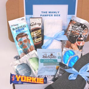 May include: A gift box labeled "THE MANLY PAMPER BOX" filled with skincare products, snacks, and accessories. Items include a face mask, eye patches, socks, a towel, and a chocolate bar. The box is tied with a blue ribbon.