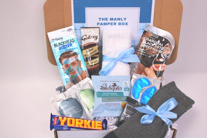 May include: A gift box labeled "THE MANLY PAMPER BOX" filled with skincare products, snacks, and accessories. Items include a face mask, eye patches, socks, a towel, and a chocolate bar. The box is tied with a blue ribbon.