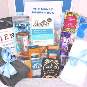 May include: A gift box labeled "THE MANLY PAMPER BOX" filled with self-care items. Includes a face mask, popcorn, chocolate, eye mask, bath tea, and a towel tied with a blue ribbon. Also includes a pair of gray washcloths tied with a blue ribbon.
