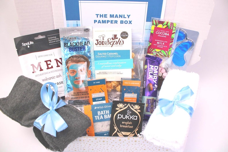 May include: A gift box labeled "THE MANLY PAMPER BOX" filled with self-care items. Includes a face mask, popcorn, chocolate, eye mask, bath tea, and a towel tied with a blue ribbon. Also includes a pair of gray washcloths tied with a blue ribbon.