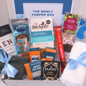 May include: A gift box labeled "THE MANLY PAMPER BOX" filled with self-care items. Includes a face mask, popcorn, chocolate, tea, soap, eye mask, socks, and a white towel tied with a blue ribbon.