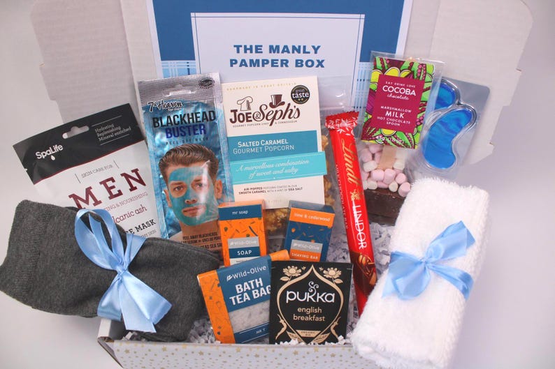 May include: A gift box labeled "THE MANLY PAMPER BOX" filled with self-care items. Includes a face mask, popcorn, chocolate, tea, soap, eye mask, socks, and a white towel tied with a blue ribbon.