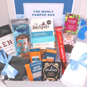 May include: A gift box labeled "THE MANLY PAMPER BOX" filled with self-care items. Includes a face mask, popcorn, soap, tea, chocolate, eye mask, and a gray sock tied with a blue ribbon. The box is white with a blue accent.