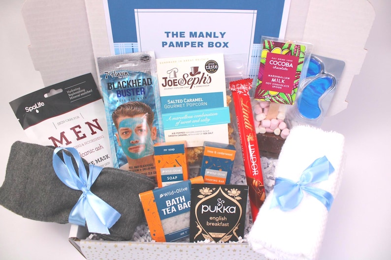 May include: A gift box labeled "THE MANLY PAMPER BOX" filled with self-care items. Includes a face mask, popcorn, soap, tea, chocolate, eye mask, and a gray sock tied with a blue ribbon. The box is white with a blue accent.