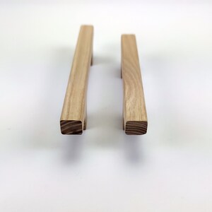 Puede incluir: Dos tiradores de madera para armarios con acabado natural. Cada tirador es rectangular con base cuadrada y asa ligeramente curvada. La veta de la madera es visible. Dise&ntilde;ado para muebles.