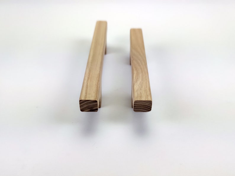 Puede incluir: Dos tiradores de madera para armarios con acabado natural. Cada tirador es rectangular con base cuadrada y asa ligeramente curvada. La veta de la madera es visible. Dise&ntilde;ado para muebles.