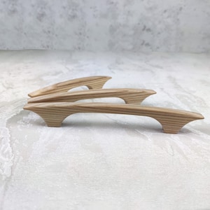 May include: Four wooden cabinet pulls with a natural wood grain finish. The pulls have a curved design with flared ends, and are arranged on a light-coloured surface. These are likely for kitchen or bathroom cabinets.