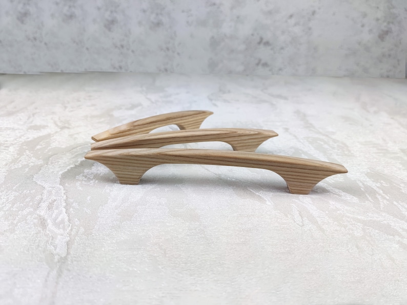 May include: Four wooden cabinet pulls with a natural wood grain finish. The pulls have a curved design with flared ends, and are arranged on a light-coloured surface. These are likely for kitchen or bathroom cabinets.