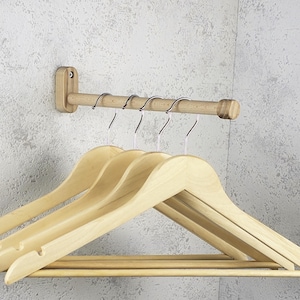 Puede incluir: Un perchero de pared de madera con una barra horizontal y cinco ganchos metálicos. Debajo del perchero hay varias perchas de madera de color claro. El perchero está fijado a una pared gris texturizada.