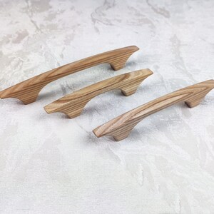 May include: Three wooden cabinet pulls with a natural wood grain finish. The pulls have a curved, ergonomic design and are arranged diagonally. The wood has a light brown colour with darker grain patterns, suggesting a natural, unfinished look.