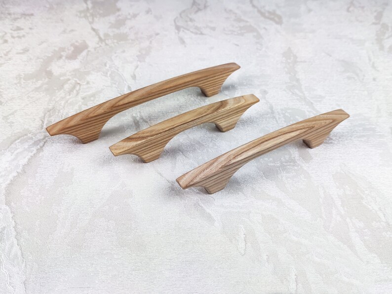 May include: Three wooden cabinet pulls with a natural wood grain finish. The pulls have a curved, ergonomic design and are arranged diagonally. The wood has a light brown colour with darker grain patterns, suggesting a natural, unfinished look.