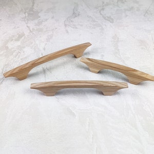 May include: Three wooden cabinet handles with a natural wood grain finish. The handles have a curved design with a decorative base. Suitable for drawers and cabinets. Each handle is approximately 15 cm long.