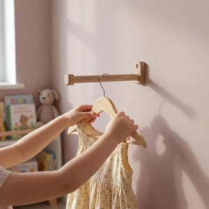 May include: A light-coloured wooden wall-mounted rod with a cylindrical end cap, featuring a matching wooden clothes hanger suspended from the rod. The hanger holds a cream-coloured sleeveless garment with a small brown floral pattern throughout.