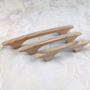 May include: Four wooden cabinet handles of different sizes. The handles are made of light-coloured wood with visible grain and a curved design. They are arranged on a white surface.