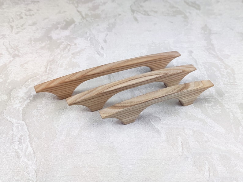 May include: Four wooden cabinet handles of different sizes. The handles are made of light-coloured wood with visible grain and a curved design. They are arranged on a white surface.