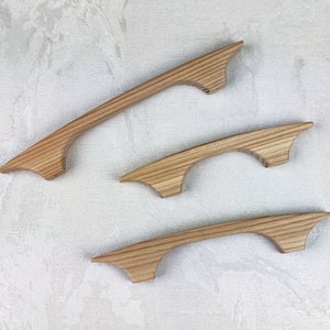 May include: Three wooden cabinet handles with a natural finish. The handles have a curved design with a flared end. The wood grain is visible, adding texture. Suitable for kitchen or bathroom cabinets.