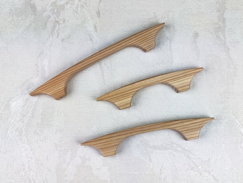 May include: Three wooden cabinet handles with a natural finish. The handles have a curved design with a flared end. The wood grain is visible, adding texture. Suitable for kitchen or bathroom cabinets.