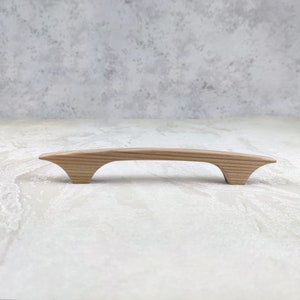 May include: A light-coloured wooden cabinet pull with a curved handle and flared ends. The hardware is made of natural wood and is designed for easy gripping. The cabinet pull is approximately 18 cm long.