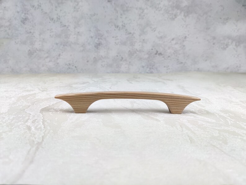May include: A light-coloured wooden cabinet pull with a curved handle and flared ends. The hardware is made of natural wood and is designed for easy gripping. The cabinet pull is approximately 18 cm long.