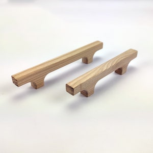 Puede incluir: Dos tiradores de armario de madera con acabado natural. Cada tirador tiene una barra rectangular con bordes redondeados y dos peque&ntilde;os pies cuadrados. La veta de la madera es visible. Los tiradores est&aacute;n dise&ntilde;ados para muebles.