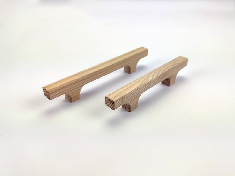 Puede incluir: Dos tiradores de armario de madera con acabado natural. Cada tirador tiene una barra rectangular con bordes redondeados y dos peque&ntilde;os pies cuadrados. La veta de la madera es visible. Los tiradores est&aacute;n dise&ntilde;ados para muebles.