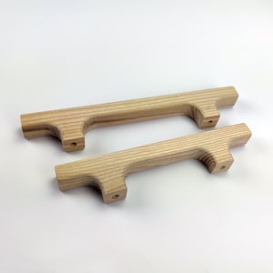 Puede incluir: Dos tiradores de armario de madera clara natural. Cada tirador tiene un cuerpo rectangular con un asa curvada y dos orificios pretaladrados para el montaje. Los tiradores son de diferentes tama&ntilde;os.