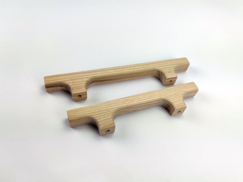 Puede incluir: Dos tiradores de armario de madera clara natural. Cada tirador tiene un cuerpo rectangular con un asa curvada y dos orificios pretaladrados para el montaje. Los tiradores son de diferentes tama&ntilde;os.