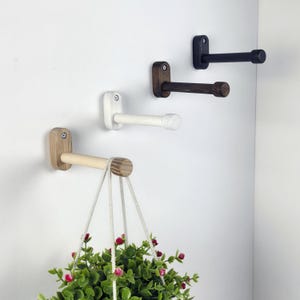May include: Four wooden wall hooks in natural, white, dark brown, and black. Each hook has a rectangular base and a cylindrical arm. One hook supports a hanging plant with green foliage and red flowers.