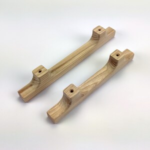 Puede incluir: Dos tiradores de armario de madera clara con veta natural. Los tiradores tienen un dise&ntilde;o curvo con extremos cuadrados y agujeros pretaladrados para el montaje. Los tiradores miden unos 25 cm de largo.