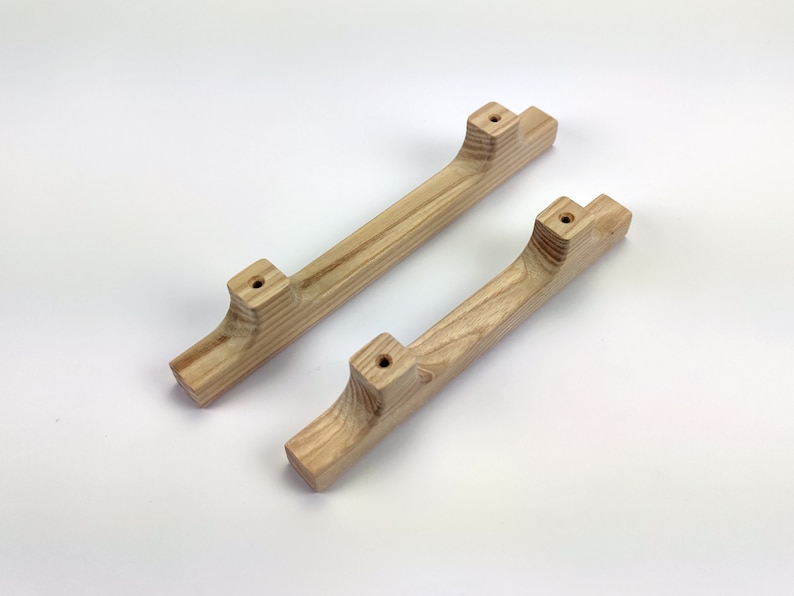 Puede incluir: Dos tiradores de armario de madera clara con veta natural. Los tiradores tienen un dise&ntilde;o curvo con extremos cuadrados y agujeros pretaladrados para el montaje. Los tiradores miden unos 25 cm de largo.