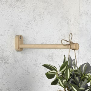 May include: A light-colored wooden plant hanger mounted on a textured wall. The hanger has a cylindrical rod with a decorative end and a jute twine bow. A hanging plant with green and white striped leaves is suspended from the hanger.
